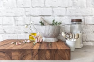 Thick Wild Wood Mogo Chop Cutting Carving Serving Board with garlic, rosemary, olive oil, and a mortar and pestle, displayed in a rustic kitchen setting with utensils and a white brick wall in the background.