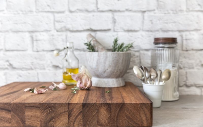 Thick Wild Wood Mogo Chop Cutting Carving Serving Board with garlic, rosemary, olive oil, and a mortar and pestle, displayed in a rustic kitchen setting with utensils and a white brick wall in the background.