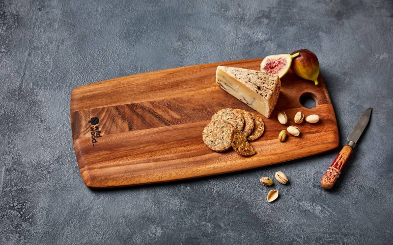 Wooden serving board with cheese, crackers, figs and pistachios on a grey surface, Wild Wood logo visible.